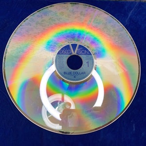 May include: A vintage Discovision disc with a rainbow-like iridescent sheen. The disc has a blue label with the text "BLUE COLLAR" and "DISCOVISION." The disc is circular and reflects light, creating a colorful effect.