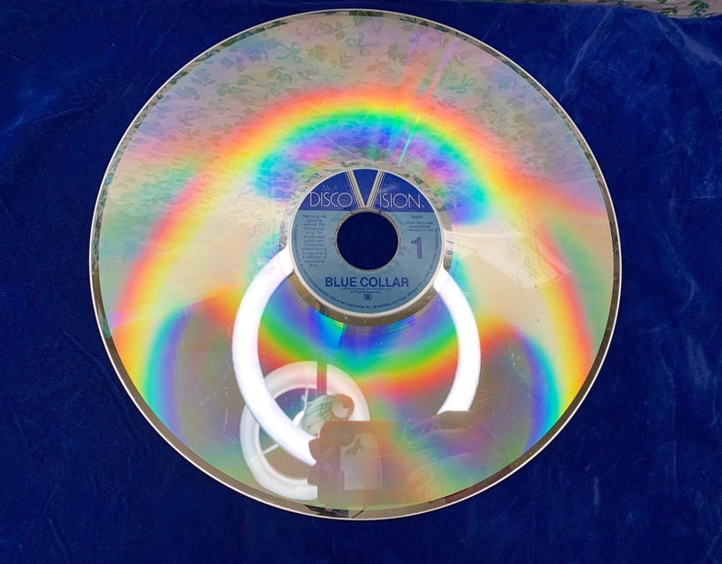 May include: A vintage Discovision disc with a rainbow-like iridescent sheen. The disc has a blue label with the text "BLUE COLLAR" and "DISCOVISION." The disc is circular and reflects light, creating a colorful effect.