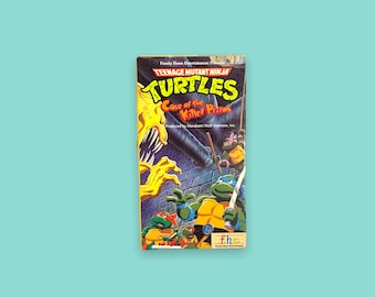 Pode incluir: Uma fita VHS de "Teenage Mutant Ninja Turtles: Case of the Killer Pizzas". A capa apresenta as Tartarugas Ninja, um monstro amarelo e o texto do título. A fita está em um fundo azul claro.