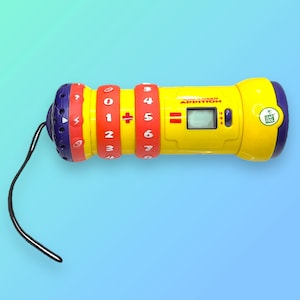 Può includere: Un giocattolo educativo LeapFrog giallo e viola con un display digitale e anelli numerici rotanti. Il giocattolo presenta le parole "Smart Addition" e un logo LeapFrog. Un cavo nero è attaccato all'estremità.