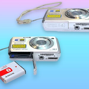 May include: Two silver Sony Cyber-shot digital cameras with attached wrist straps. One camera has its battery compartment open, revealing a red and white battery labeled "SONY". The cameras have a 12.1 megapixel resolution.