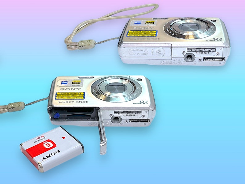 May include: Two silver Sony Cyber-shot digital cameras with attached wrist straps. One camera has its battery compartment open, revealing a red and white battery labeled "SONY". The cameras have a 12.1 megapixel resolution.