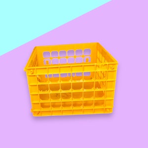 Puede incluir: Una caja de plástico amarilla brillante con forma rectangular y un patrón de rejilla. La caja tiene múltiples aberturas y está diseñada para almacenamiento o transporte. El fondo es una división de azul claro y morado.
