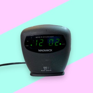 May include: A black Magnavox AJ3145 AM/FM clock radio with a green digital display showing the time as 12:02. The clock has a dual alarm feature and a power cord.