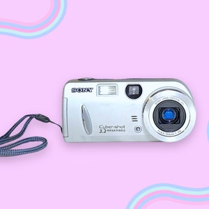 May include: Silver Sony Cybershot 3.2 Megapixel digital camera with a blue and gray wrist strap. The camera has a flash, lens, and various buttons. The background is pink with pastel colored wavy lines.