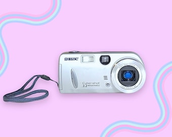 Pode incluir: Câmera digital Sony Cybershot prateada de 3,2 megapixels com uma alça de pulso azul e cinza. A câmera possui flash, lente e vários botões. O fundo é rosa com linhas onduladas em cores pastel.