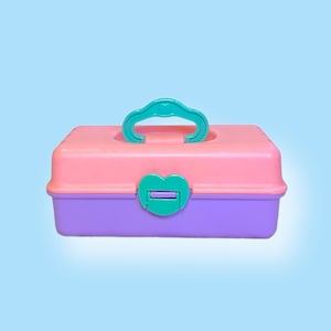 May include: A vintage, rectangular, plastic storage box in pastel colors. The top is pink, the bottom is purple, and the handle and clasp are teal. The box has a rounded top and a heart-shaped clasp.