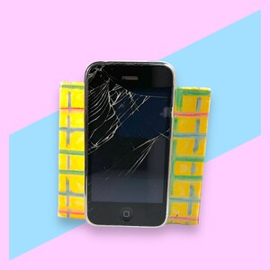 May include: A black smartphone with a cracked screen is positioned between two yellow and green rectangular blocks. The background features a pink surface with a blue diagonal shape.