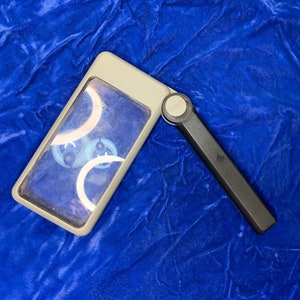 May include: A gray rectangular magnifying glass with a black handle, open and angled. The lens shows a design with white and blue curved shapes. The magnifying glass is on a blue velvet surface.