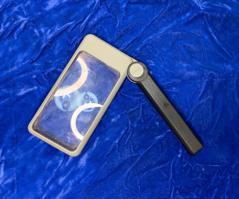 May include: A gray rectangular magnifying glass with a black handle, open and angled. The lens shows a design with white and blue curved shapes. The magnifying glass is on a blue velvet surface.