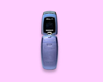 May include: A blue Samsung flip phone, open to reveal the screen. The phone has a Verizon logo and the Samsung brand name is visible. The phone is set against a pink background.
