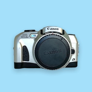 Pode incluir: C&acirc;mera Canon EOS IX Lite prateada com tampa de lente preta. O logotipo da Canon est&aacute; na parte superior e na tampa da lente. O nome do modelo "EOS IX Lite" &eacute; vis&iacute;vel no corpo da c&acirc;mera. Em um fundo azul claro.