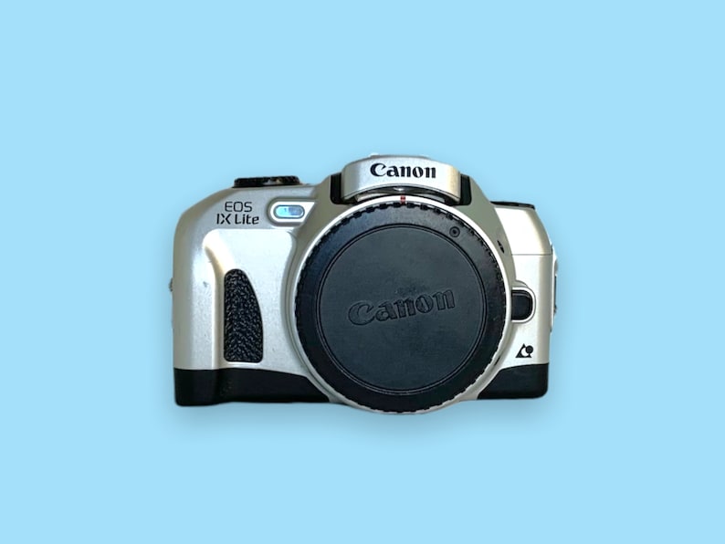 Pode incluir: C&acirc;mera Canon EOS IX Lite prateada com tampa de lente preta. O logotipo da Canon est&aacute; na parte superior e na tampa da lente. O nome do modelo "EOS IX Lite" &eacute; vis&iacute;vel no corpo da c&acirc;mera. Em um fundo azul claro.