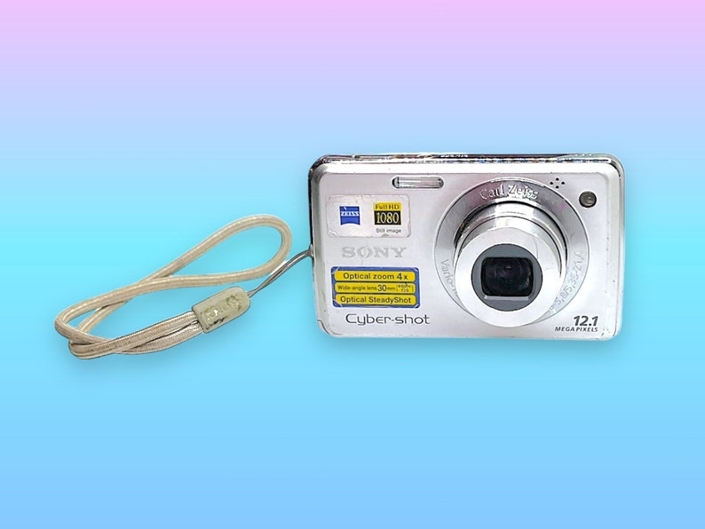 May include: Silver Sony Cyber-shot digital camera with a Carl Zeiss lens and a 12.1 megapixel resolution. Features include optical zoom and optical steady shot. Includes a beige wrist strap.