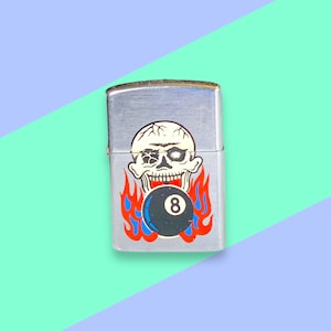 May include: A silver metal lighter featuring a skull and flaming eight ball design. The skull is white with black detailing, and the flames are red and blue. The rectangular lighter has a hinged lid.