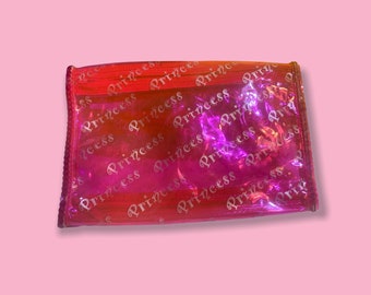 May include: A rectangular, iridescent pink and red pouch with the word "Princess" repeated across the surface. The pouch has a clear, transparent appearance and a zipper closure.