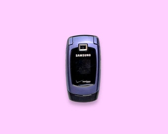 May include: A closed, blue Samsung flip phone with a Verizon logo. The phone has a camera and a small screen. The Samsung logo is visible. The phone is on a pink background.
