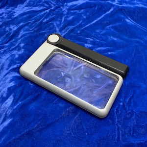 May include: A rectangular magnifying glass with a white and black handle. The magnifying glass has a clear lens and a white frame. The handle is black and has a small white button. The item is on a blue surface.