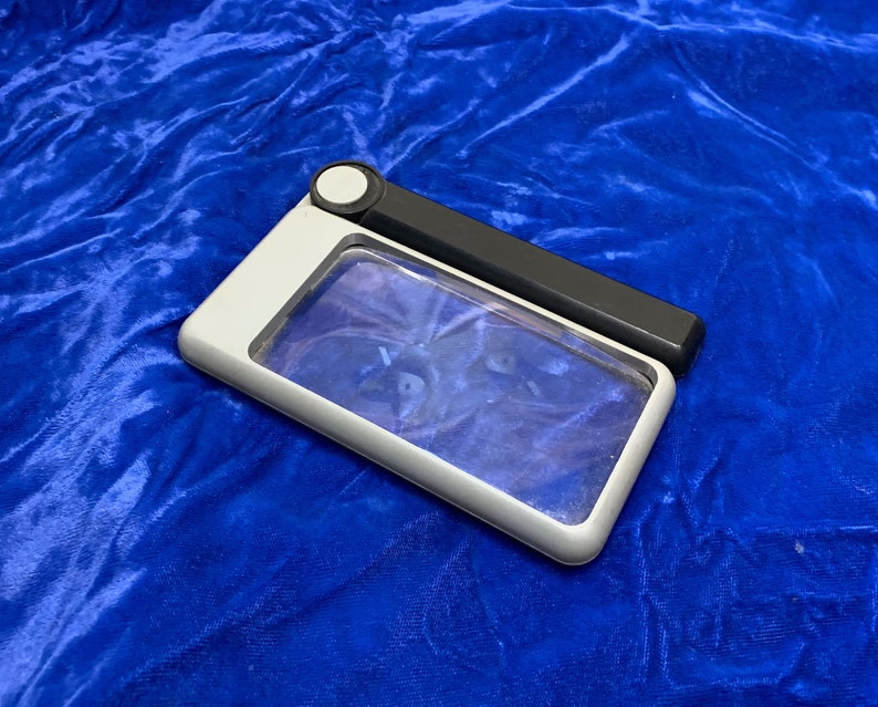 May include: A rectangular magnifying glass with a white and black handle. The magnifying glass has a clear lens and a white frame. The handle is black and has a small white button. The item is on a blue surface.
