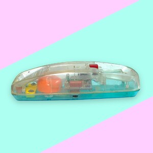 K&ouml;nnte beinhalten: Ein Vintage-Spielzeugtelefon aus klarem Kunststoff mit einem t&uuml;rkisfarbenen Sockel und einem roten Knopf. Die inneren Komponenten, darunter eine orangefarbene Kugel und gelbe Teile, sind durch das transparente Geh&auml;use sichtbar. Das Spielzeug befindet sich vor einem pastellfarbenen rosa und blauen Hintergrund.