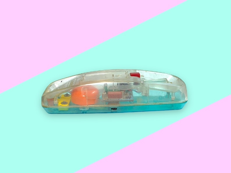 K&ouml;nnte beinhalten: Ein Vintage-Spielzeugtelefon aus klarem Kunststoff mit einem t&uuml;rkisfarbenen Sockel und einem roten Knopf. Die inneren Komponenten, darunter eine orangefarbene Kugel und gelbe Teile, sind durch das transparente Geh&auml;use sichtbar. Das Spielzeug befindet sich vor einem pastellfarbenen rosa und blauen Hintergrund.
