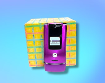 May include: A purple Motorola RAZR cell phone with a black screen displaying "Insert SIM" is positioned in front of a yellow box with a colorful grid pattern. The phone has the Motorola logo.