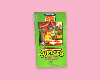 May include: A green Burger King Kids Club VHS tape featuring Teenage Mutant Ninja Turtles: Invasion of the Turtle Snatchers. The tape has colorful illustrations of the turtles and a woman. The Burger King logo is at the top.