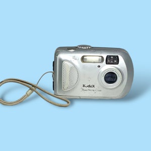 Könnte beinhalten: Silberne Kodak EasyShare C300 Digitalkamera mit beigefarbenem Handgelenkband. Die Kamera hat einen Blitz, ein Objektiv und verschiedene Knöpfe. Der Text "Kodak EasyShare C300" ist aufgedruckt.