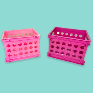 Puede incluir: Dos cajas de almacenamiento de plástico rosa con forma rectangular y agujeros redondos. Las cajas tienen un asa en cada extremo y están sobre un fondo azul claro. Las cajas están hechas de un material plástico duradero.