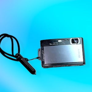 May include: A silver and black Sony Cybershot digital camera with a black wrist strap. The camera has a lens, flash, and various buttons. The text "Cybershot" is visible on the side.