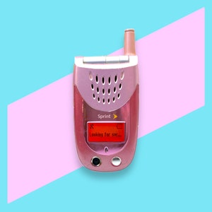 Könnte beinhalten: Ein pinkfarbenes Sprint-Klapphandy mit einem kleinen Bildschirm, auf dem "Looking for SVC..." steht. Das Telefon hat eine silberne Antenne und Lautsprecherlöcher. Der Hintergrund ist ein hellblaues und rosa geometrisches Design.