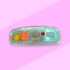 K&ouml;nnte beinhalten: Ein Vintage-Spielzeugtelefon aus transparentem Kunststoff, das seine inneren Komponenten zeigt. Das Telefon hat ein leuchtend orangefarbenes Ziffernblatt, einen gelben H&ouml;rer und einen roten Knopf. Der Hintergrund ist rosa.