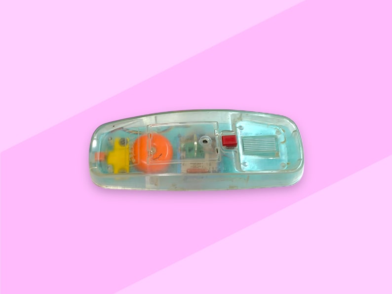 K&ouml;nnte beinhalten: Ein Vintage-Spielzeugtelefon aus transparentem Kunststoff, das seine inneren Komponenten zeigt. Das Telefon hat ein leuchtend orangefarbenes Ziffernblatt, einen gelben H&ouml;rer und einen roten Knopf. Der Hintergrund ist rosa.