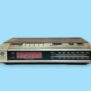 Puede incluir: Un radio despertador digital General Electric vintage con una parte superior de vetas de madera y adornos plateados. La pantalla digital roja muestra la hora como 12:02. El reloj tiene capacidades de radio AM/FM.