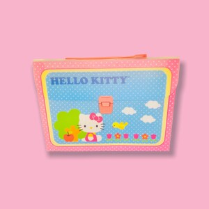 May include: A Hello Kitty lunchbox with a pink polka dot border and a blue background with white polka dots. The lunchbox features Hello Kitty, a tree, flowers, and the words "HELLO KITTY".