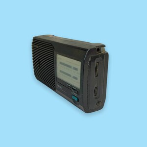 Może przedstawiać: Czarno-białe przenośne radio w stylu vintage z tunerem FM/AM. Radio ma prostokątny kształt z kratką głośnika z przodu i wyświetlaczem. Posiada przyciski i pokrętła do strojenia i regulacji głośności. Radio jest na jasnoniebieskim tle.