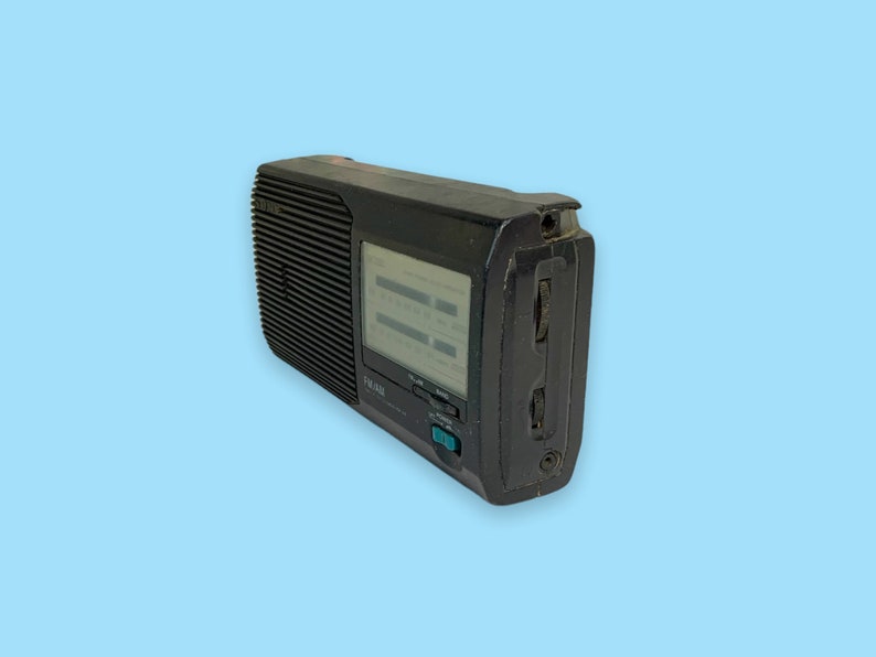 Może przedstawiać: Czarno-białe przenośne radio w stylu vintage z tunerem FM/AM. Radio ma prostokątny kształt z kratką głośnika z przodu i wyświetlaczem. Posiada przyciski i pokrętła do strojenia i regulacji głośności. Radio jest na jasnoniebieskim tle.
