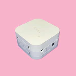 May include: A white P-touch Cube label maker on a pink background. The device is square with rounded edges and has the words "P-touch CUBE" printed on the top. It features a small circular button and a label port.