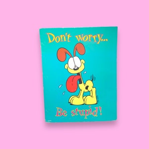 Peut inclure: Carte de couleur turquoise avec Garfield, le personnage de dessin animé, et le texte "Don't worry... Be stupid!" en jaune et rouge. Le chien est jaune avec une langue rouge et des oreilles marron.