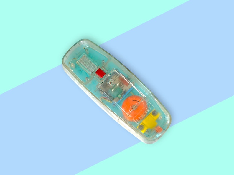 K&ouml;nnte beinhalten: Vintage Telefonh&ouml;rer aus klarem Kunststoff mit sichtbaren internen Komponenten. Der H&ouml;rer hat einen roten Knopf, einen orangefarbenen Lautsprecher und gelbe und blaue Akzente. Der Hintergrund ist hellblau und t&uuml;rkis.