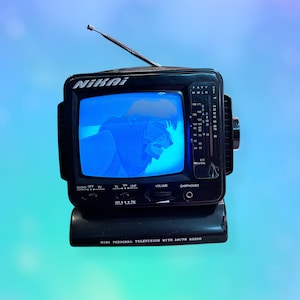 Könnte beinhalten: Kleiner, schwarzer Vintage NIKAI Mini-Personalfernseher mit AM/FM-Radio. Der Bildschirm zeigt ein blaues Bild. Der Fernseher hat eine ausziehbare Antenne und einen Drehregler für die Lautstärke. Der Text "MINI PERSONAL TELEVISION WITH AM/FM RADIO" ist auf der Basis aufgedruckt.