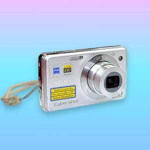 May include: Silver Sony Cyber-shot digital camera with a retractable lens. Features include Carl Zeiss lens, 4x optical zoom, and Optical SteadyShot. Includes a wrist strap. The camera has a Full HD 1080 resolution.