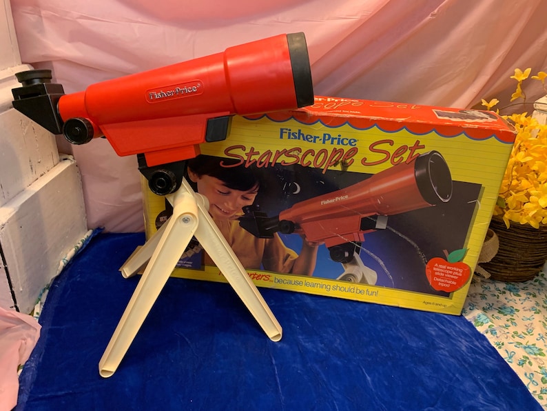 May include: A vintage Fisher-Price Starscope Set, featuring a red telescope with a black eyepiece and a cream-colored tripod. The box shows a child using the telescope. The set is designed for children.