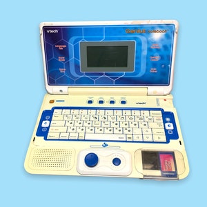 Può includere: Un computer portatile VTech Genius notebook vintage in bianco sporco e blu. Il laptop presenta un piccolo schermo, una tastiera e comandi di gioco. Il testo "Genius notebook" e "VTech" sono visibili. È inserita una cartuccia "Brain Bank".