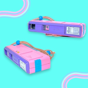 May include: A La Clic 35mm film camera featuring a bright pink and purple plastic body with a built-in flash, a viewfinder, and a tan wrist strap with a blue bead. The camera has a rectangular shape with a blue latch on the side and a yellow button.