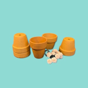 May include: A collection of terracotta flower pots in various sizes, stacked and arranged on a light blue background. A small bundle of light pink artificial roses and a string of green wire are also present. The pots are unglazed and have a classic, rustic appearance.