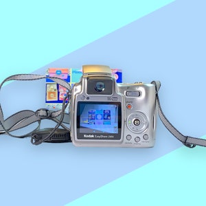 Könnte beinhalten: Silberne Kodak EasyShare Z650 Digitalkamera mit grauem Gurt. Der Bildschirm der Kamera zeigt eine farbenfrohe Benutzeroberfläche. Im Hintergrund ist ein kleines, farbenfrohes Bild zu sehen. Die Kamera hat einen Blitz und mehrere Tasten.
