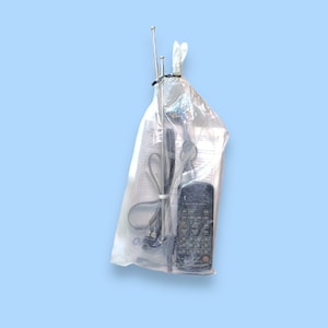 Könnte beinhalten: Eine transparente Plastiktüte mit einer schwarzen Fernbedienung, einer langen Antenne und Papieren. Die Artikel sind mit einem schwarzen Kabelbinder vor einem hellblauen Hintergrund zusammengebunden.
