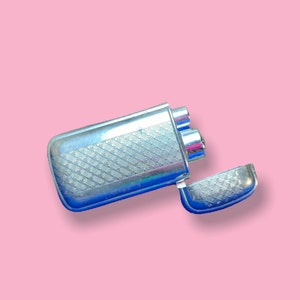 May include: A silver-colored lighter with a textured, diamond-patterned design. The lighter's lid is open, revealing the interior. The background is a solid pink color.