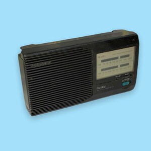 Może przedstawiać: Czarno-białe radio Sony FM/AM w stylu vintage z wyraźnym wyświetlaczem pokazującym częstotliwości radiowe. Radio ma prostokątny kształt z żebrowaną osłoną głośnika i selektorem pasma. Radio znajduje się na jasnoniebieskim tle.